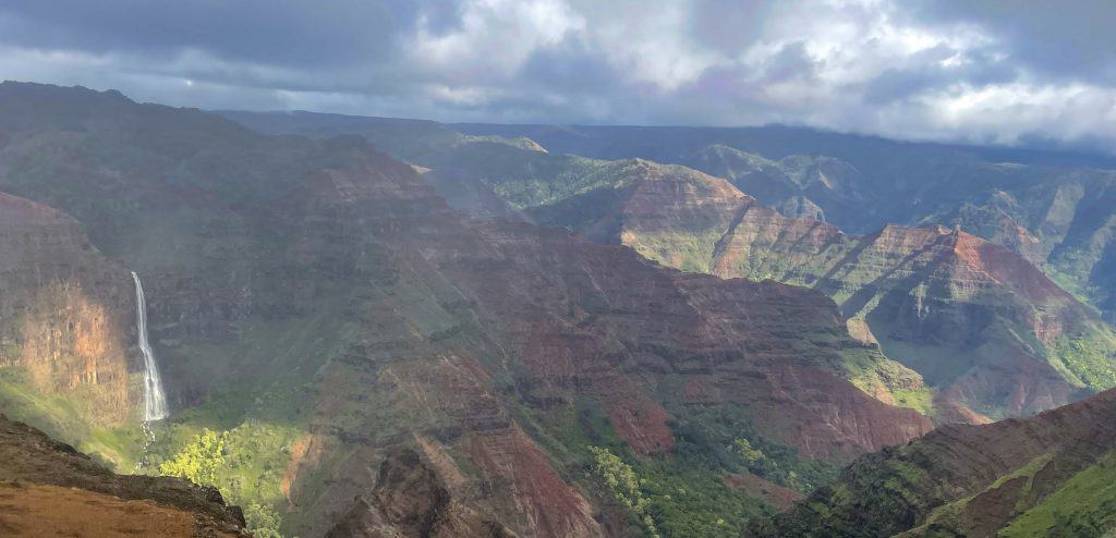 view of the Waimea Canyon with it's red cliffs, deep canyon, and tall waterfall