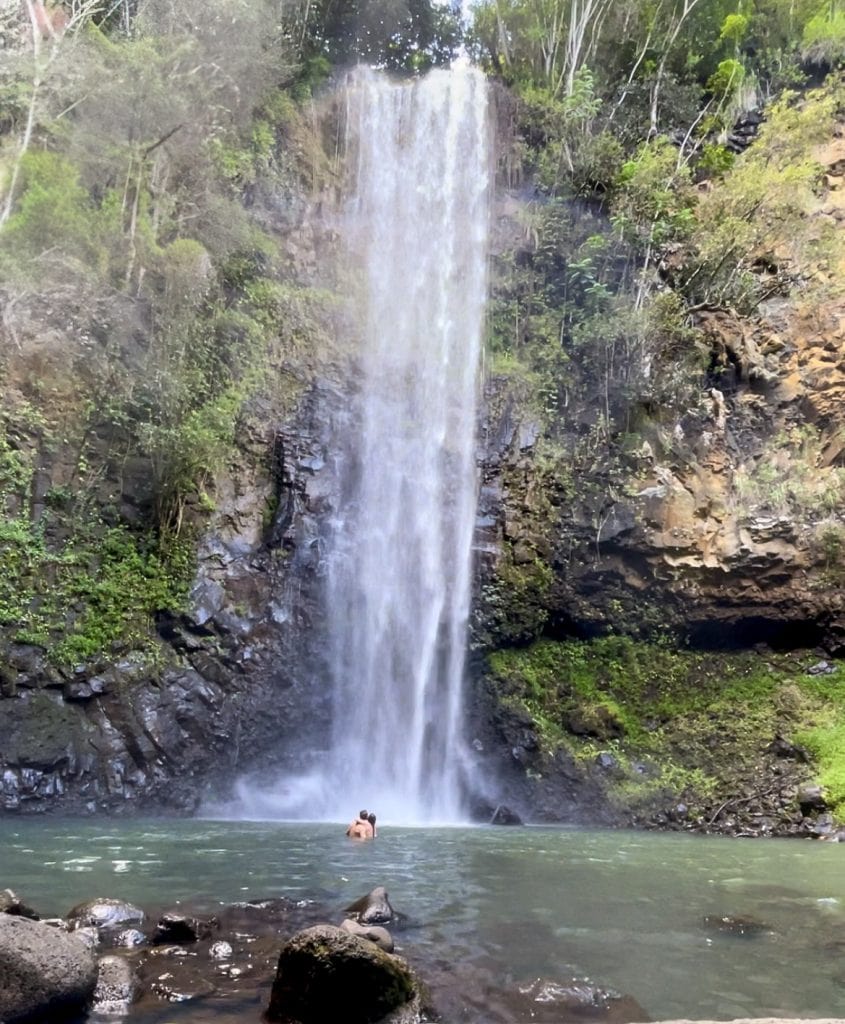 "Secret" Falls, a 100ft waterfall surrounded by dense tropical greenery. a couple is enjoying the water under the fall