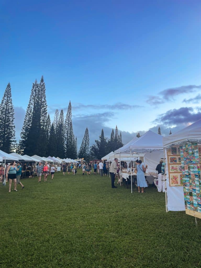 a farmers market with several white tent vendors on green grass with tall tress in the distance