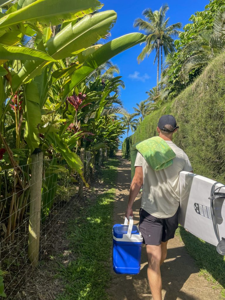 a man carrying a surfboard and a cooler through a lush alleyway to a beach in kauai