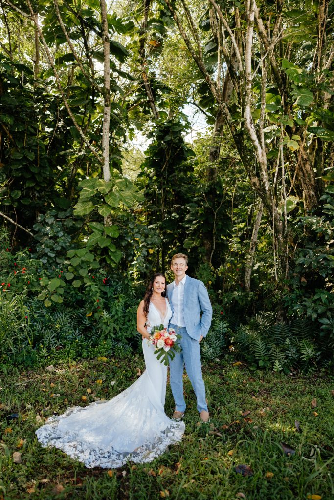 a woman in a wedding dress standing next to her new husband in a light blue suit on their elopement day in Kauai with green plants behind them
