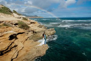 a newlywed couple hugging on sand cliffs over the ocean with a rainbow overhead