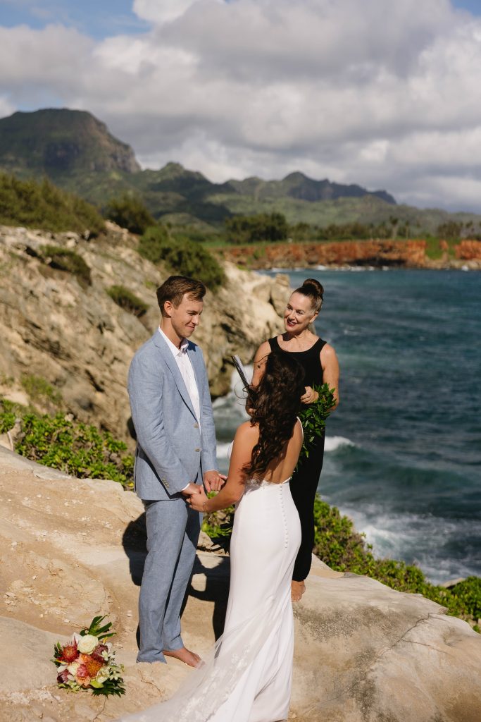 a couple elopes in Kauai with a lady officiating a ceremony on a ocean cliff with mountains in the background