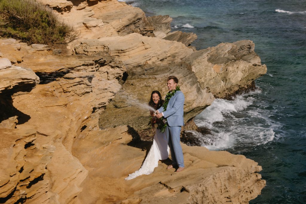 a newly married couple popping champagne on a sandy cliff in Kauai