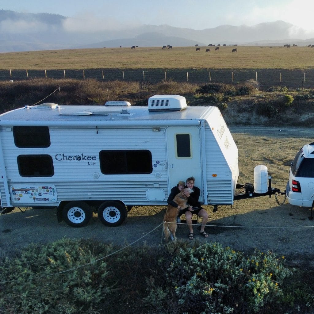 a couple and their golden retriever sitting on the steps of their small travel trailer with a scenic backdrop of golden rolling hills with cows in the field